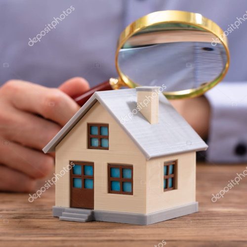 Close-up Of A Businessman's Hand Holding Magnifying Glass Over House Model Over Desk